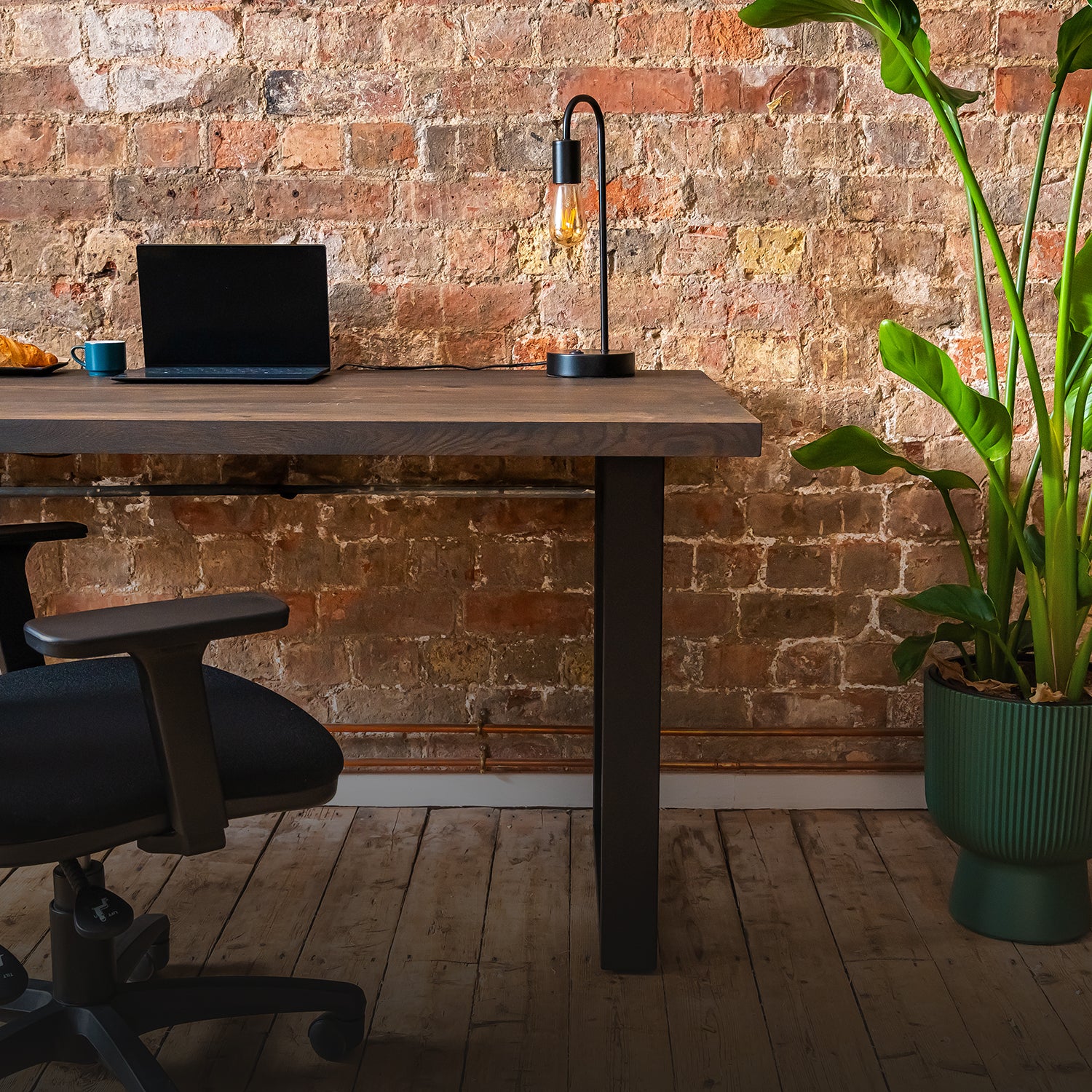 Premium oak charcoal desk with laptop, lamp, and cup against a brick wall with a plant on the right.