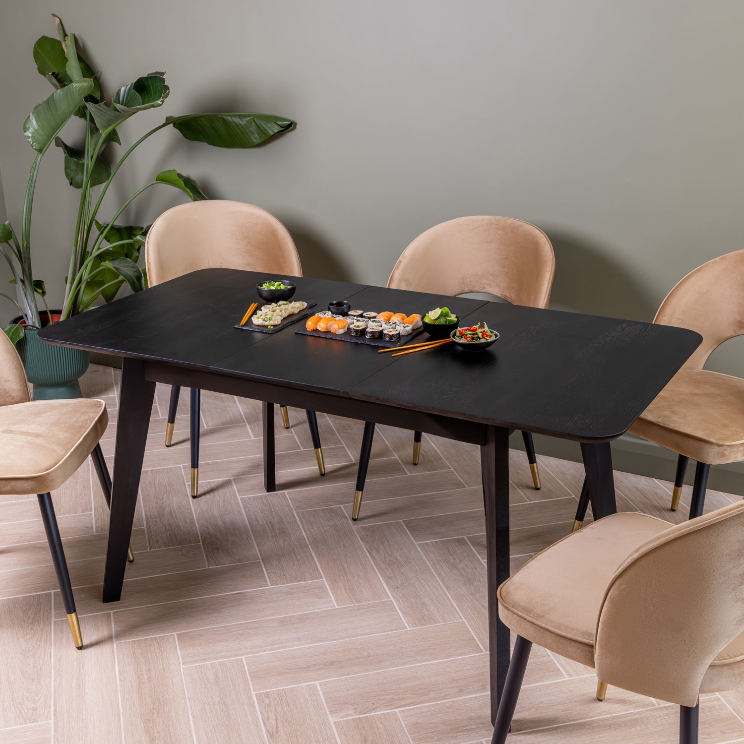 A modern dining table set in a styled room, featuring a rectangular black extendable table with five caramel upholstered velvet chairs. The table is styled with a sushi set, and the room has a green wall and light wood herringbone flooring. | 1575mm (extended)
