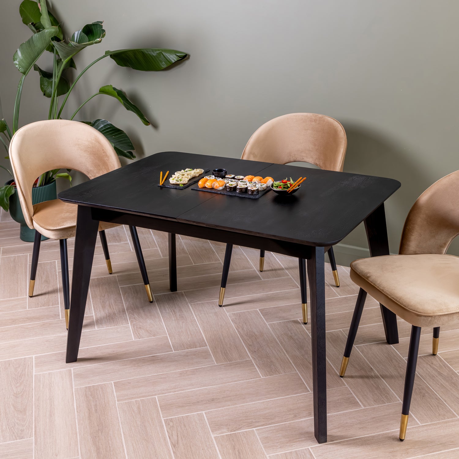 A modern dining set featuring a rectangular black extendable table and three dining chairs in caramel velvet, styled in a room with a green wall and light wood herringbone floor.