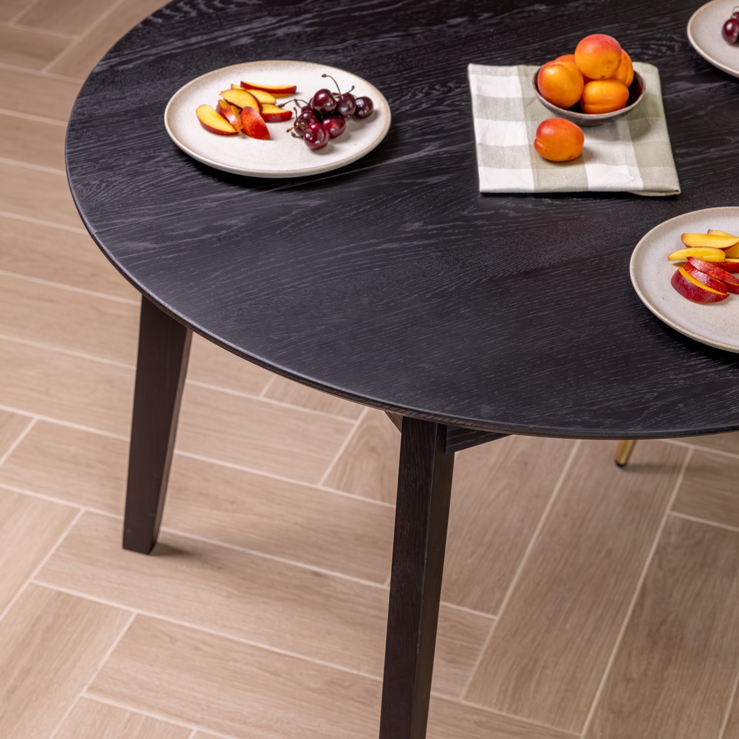 A round black wood dining table, shown in a dining room setting with light wood floor tiles. White plates with sliced apples, cherries, and an orange are styled on the table with a checkered napkin.