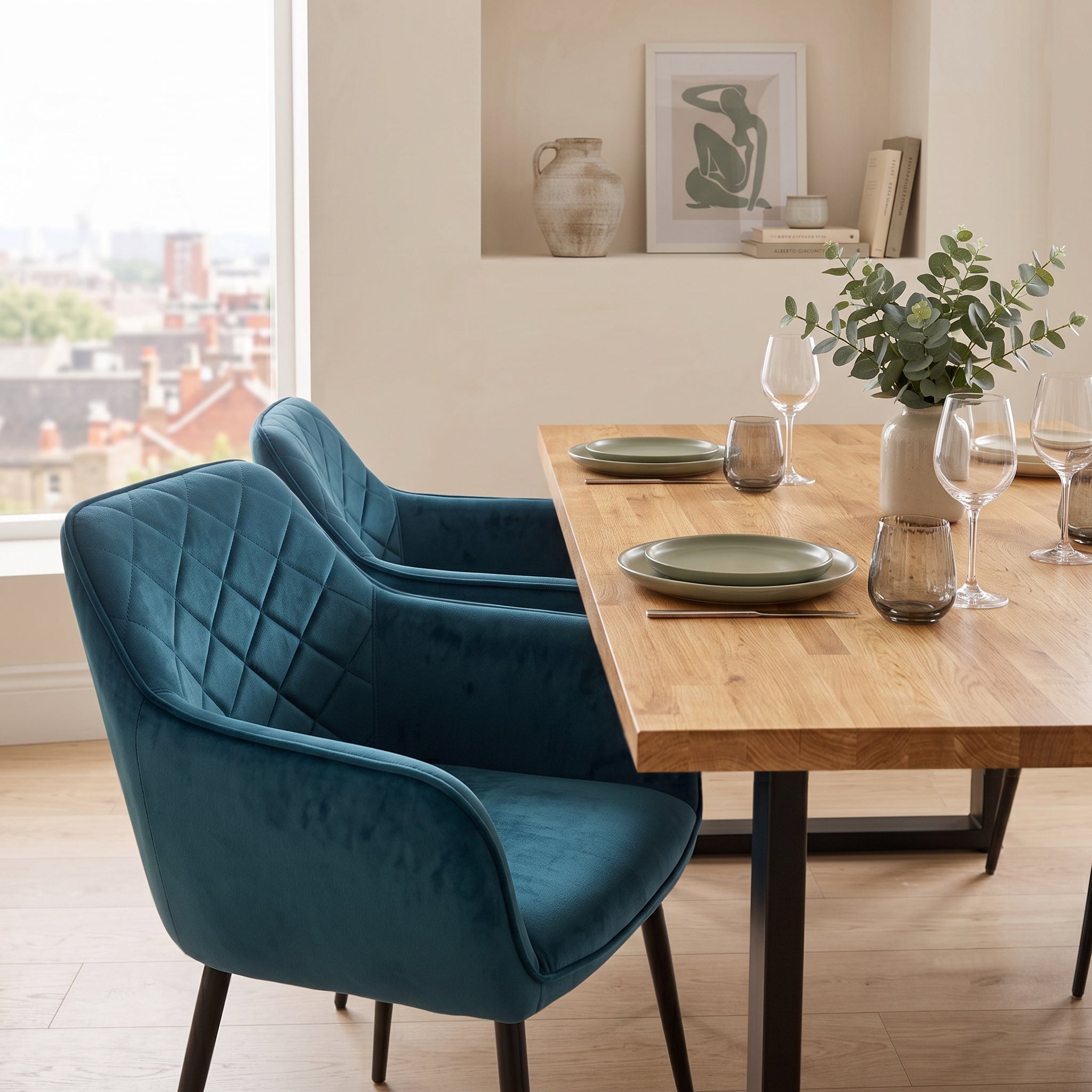 A modern dining room scene featuring two blue velvet upholstered chairs around a solid oak table set with plates and wine glasses.