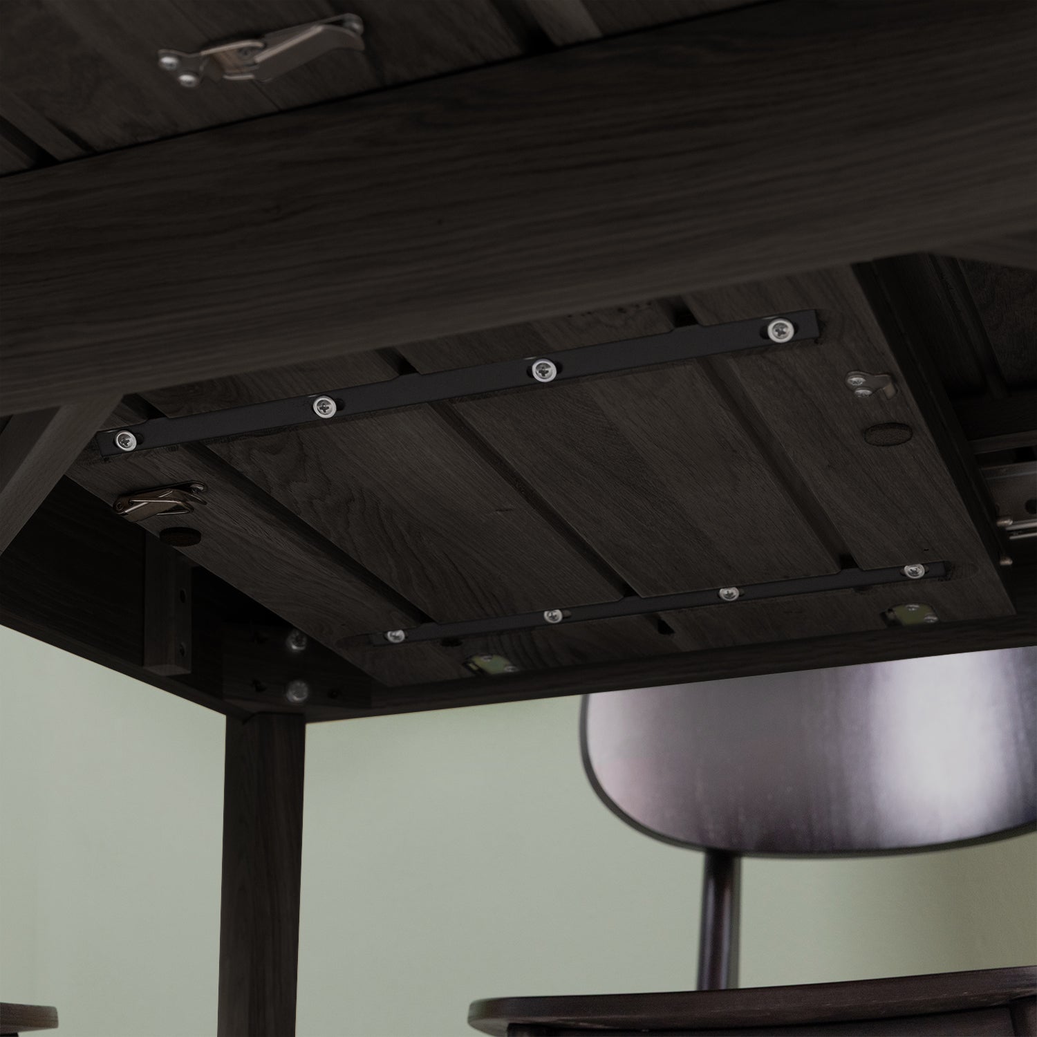 Close-up of the underside of a black wood table displaying the folding mechanism of the extendable table, with part of a chair visible in the background.