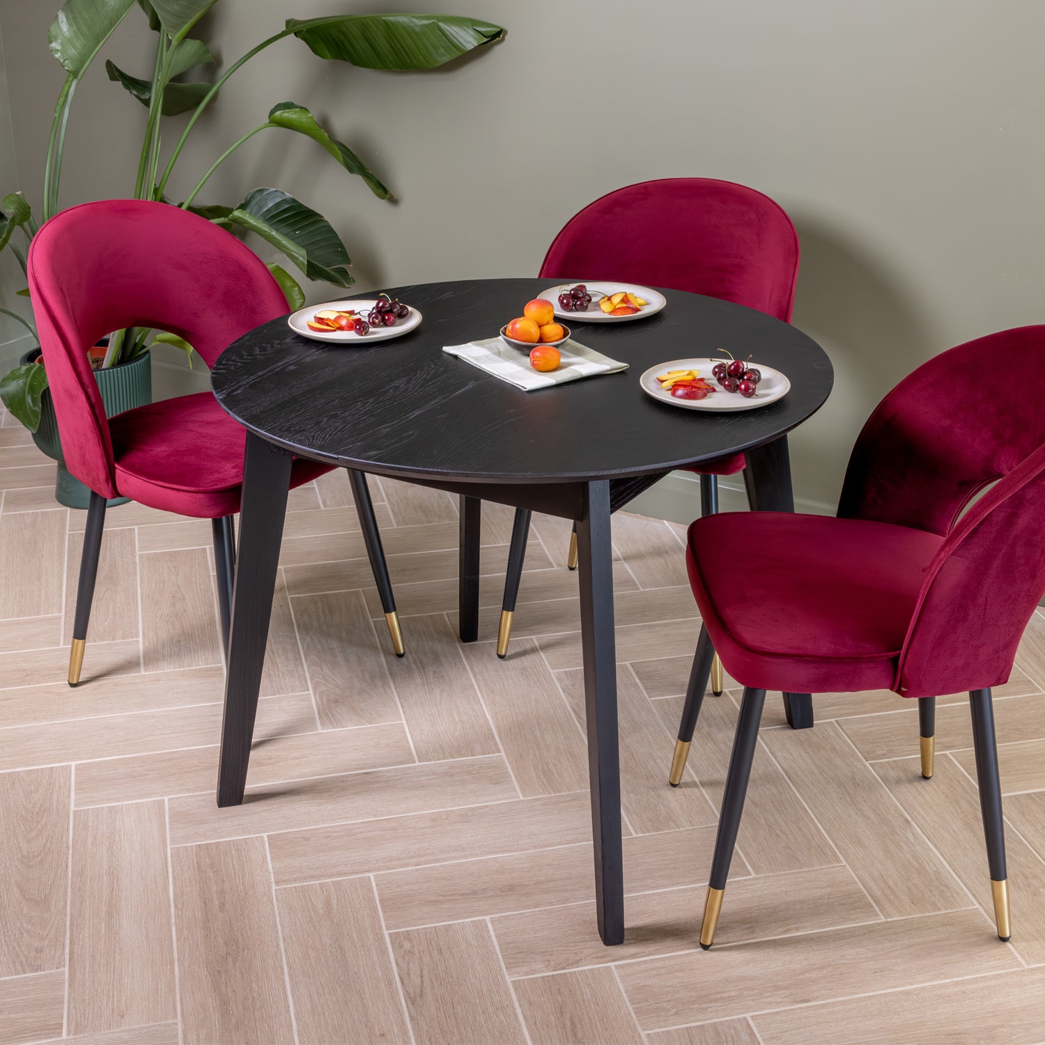 A round black dining table with four plush oxblood velvet chairs, styled in a dining room setting with a light herringbone floor, a green potted plant, and plates of fruit.