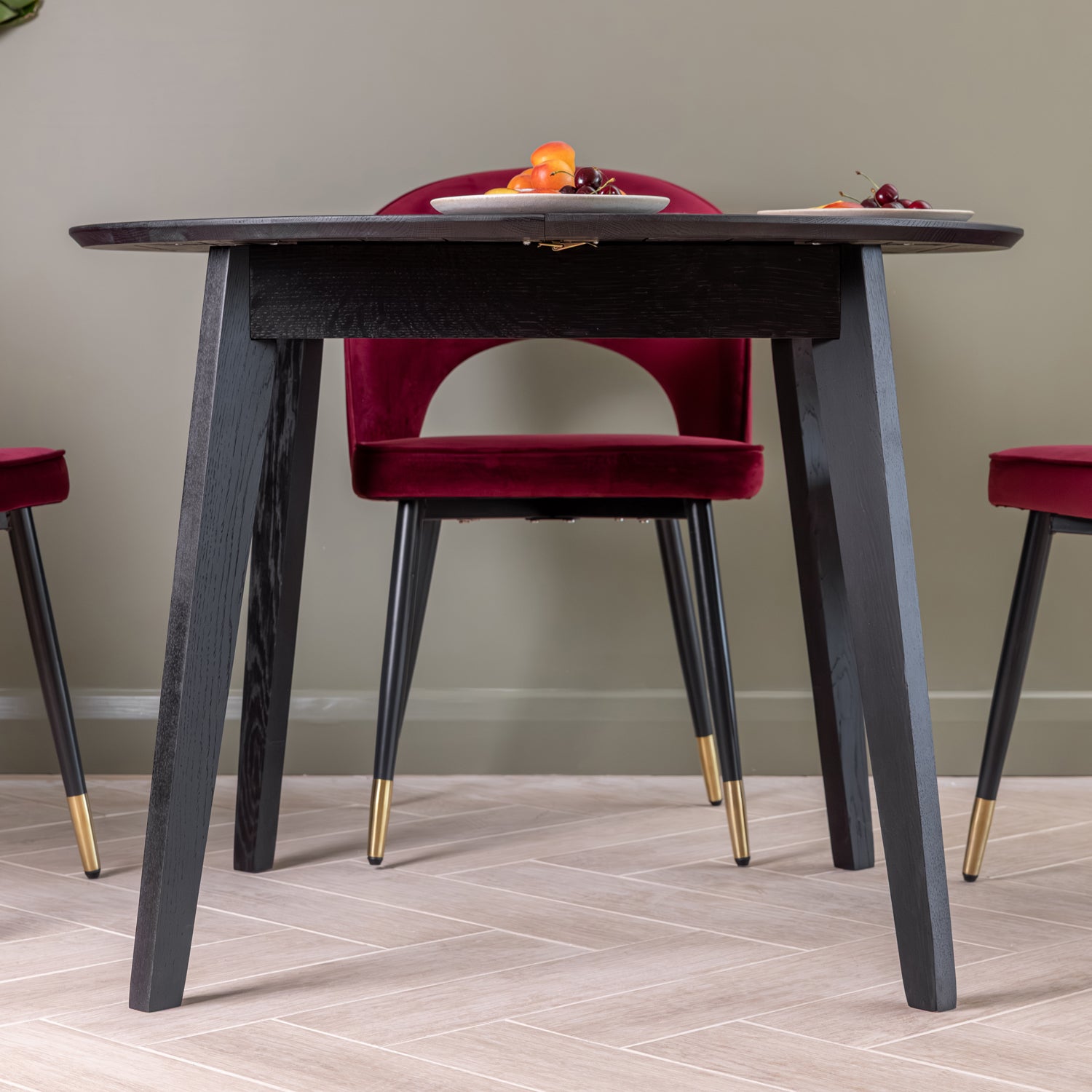 A modern extendable black dining table, paired with deep oxblood velvet chairs featuring black legs with gold champagne capped tips. The scene is set in a dining room with light wood flooring and a sage green wall.