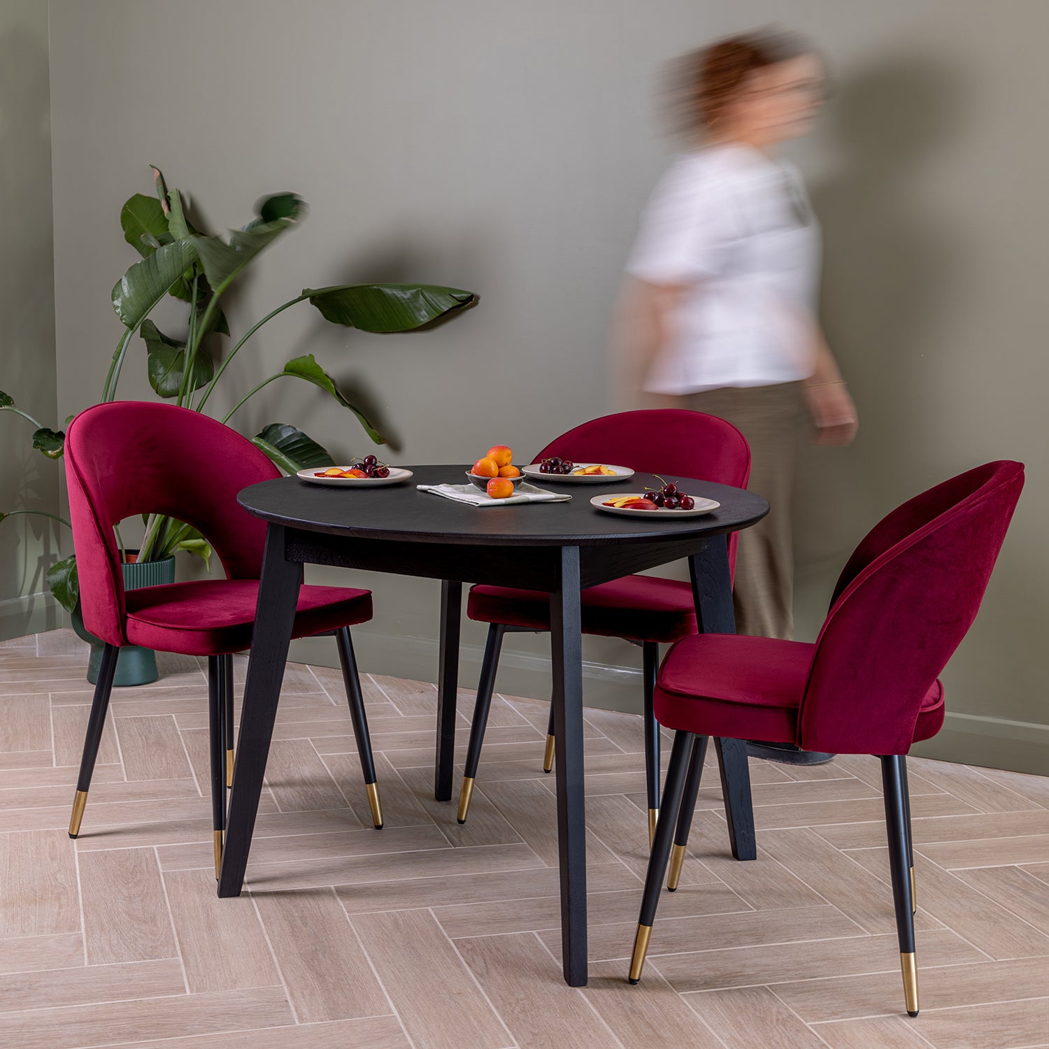 A dining set featuring a round black extendable table and three velvet chairs in oxblood, styled in a modern dining room with a blurred person walking in the background.
