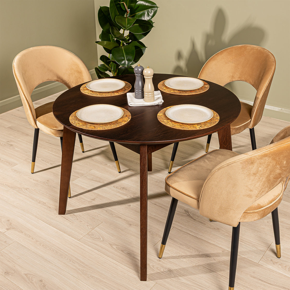 A round extendable oak dining table in walnut finish with three dining chairs in a caramel velvet fabric, styled with plates and a plant in a room with light wood floors and a sage green wall.