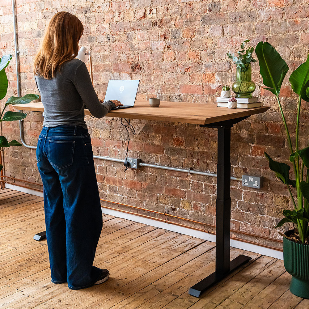 Person using a solid wood standing desk with powder-coated steel legs in a brick-walled workspace.