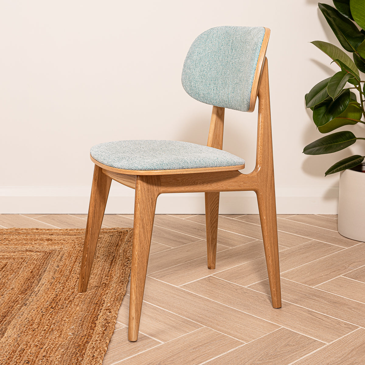 A wooden dining chair with a sea green upholstered seat and backrest, shown in a styled room setting with a woven rug, and a potted plant.