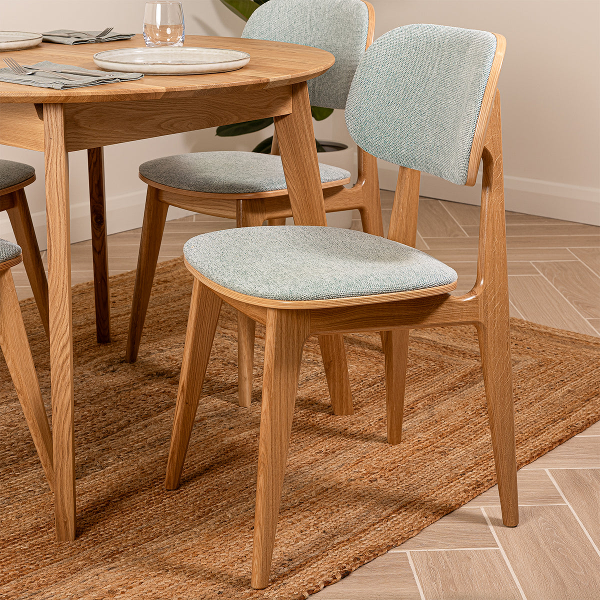 A dining set featuring a round natural oak wood table and four matching chairs with sea green upholstered seats and backs, arranged on a woven jute rug in a dining room.