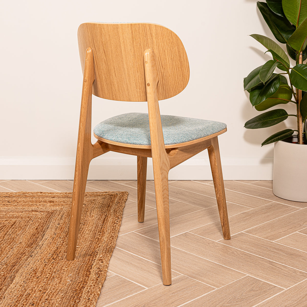 An oak wood dining chair with a curved backrest and a sea green upholstered seat cushion, shown in a styled room setting with a potted plant and woven rug,