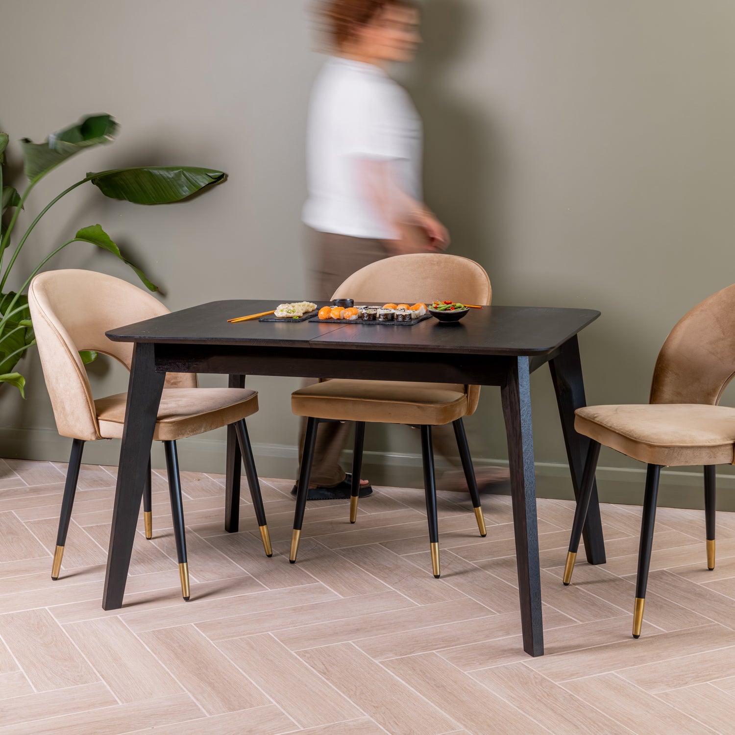 A modern dining set with a rectangular black extendable table and three upholstered velvet chairs in caramel, shown in a styled dining room with a blurred person walking in the background.