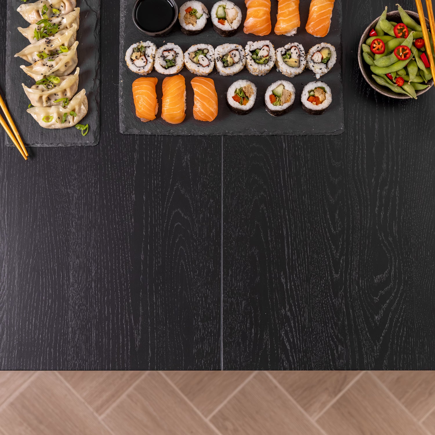A top-down view of a black oak wooden table styled with a large black slate platter of assorted sushi, including salmon nigiri, maki rolls, and a side of dumplings on another slate platter, with a bowl of edamame and red chili slices nearby.