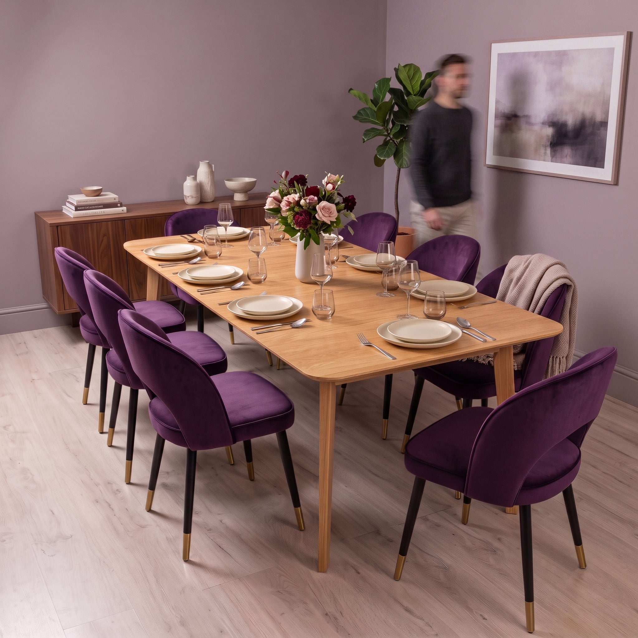A wooden dining table set with white plates and glassware, surrounded by purple velvet dining chairs with black and gold legs, in a modern dining room with a blurred man in the background.