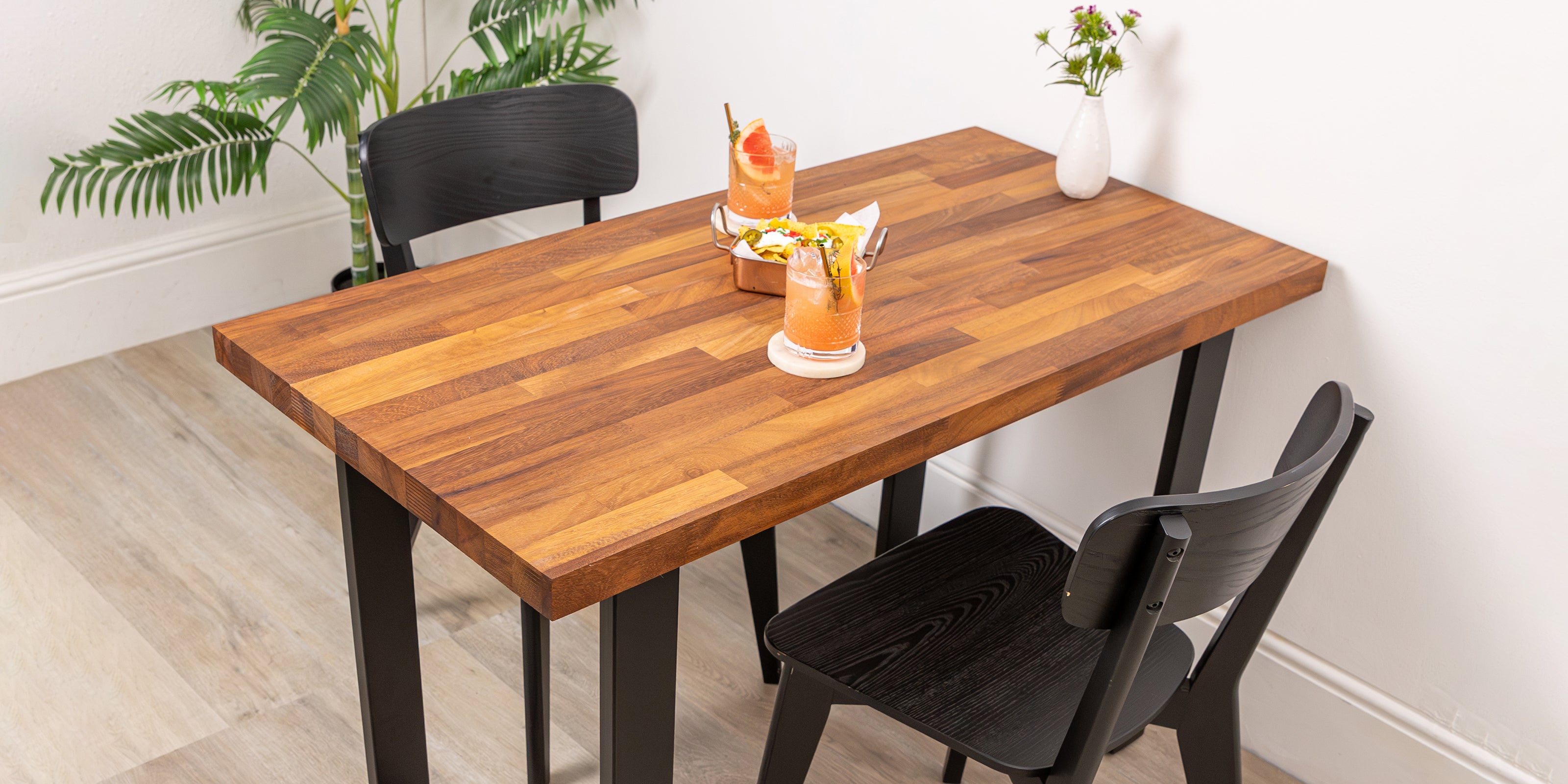 Iroko solid wood dining table with black chairs and drinks on a light wood floor.