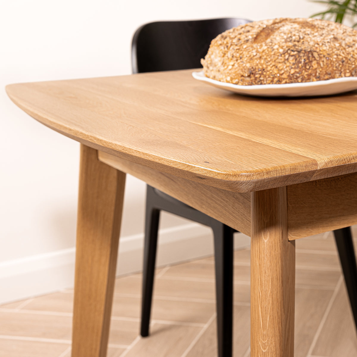 A light natural oak wooden dining table with rounded corners and tapered legs, shown in a home setting with a black chair and a seeded bread loaf on a white plate.
