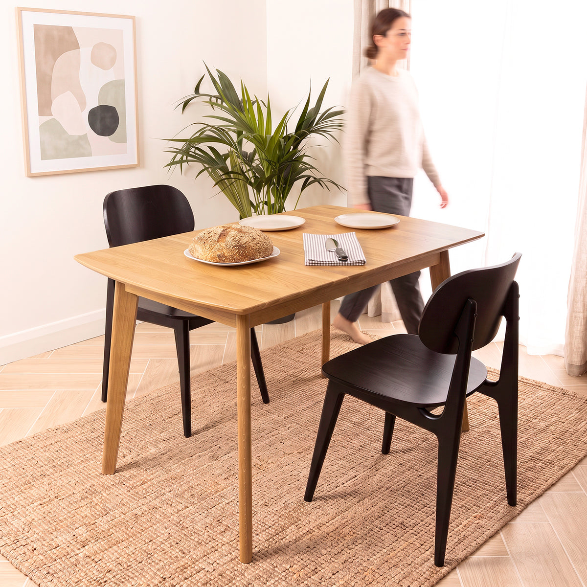 A light natural oak wood dining table with black chairs in a bright, modern dining room. A woman is blurred in the background near a window, and a potted plant and a loaf of bread are styled on the table.