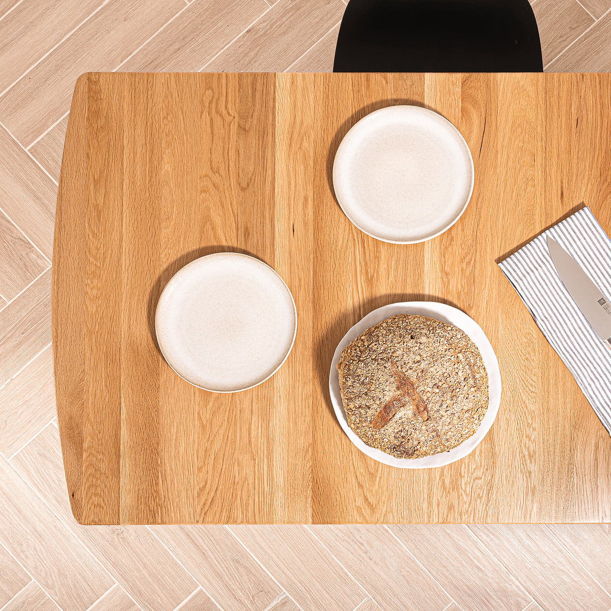 Top-down view of a rectangular Otti solid oak dining table with three white plates, one with a seeded loaf of bread and on a striped napkin, set on a light wood herringbone floor.