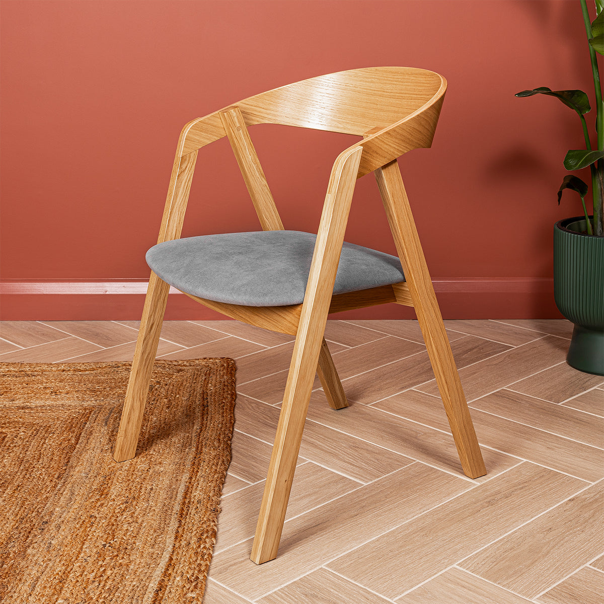 A side on view of a wooden dining chair with a curved backrest and a grey velvet seat, shown in a styled room setting with a terracotta wall, a woven rug, and a potted plant.