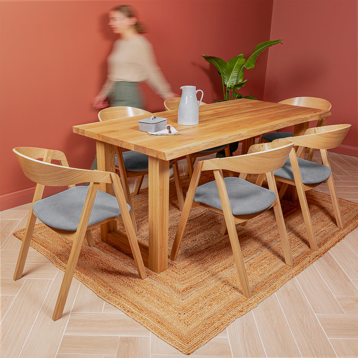 A natural wood dining table with six matching chairs featuring grey velvet seats, arranged on a woven jute rug in a dining room with terracotta-colored walls. A blurred person walks in the background near a potted plant.