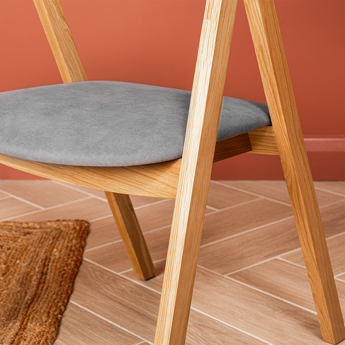 A close up of a wooden dining chair with a soft grey velvet seat, shown in a styled room setting with a terracotta wall and a woven rug.
