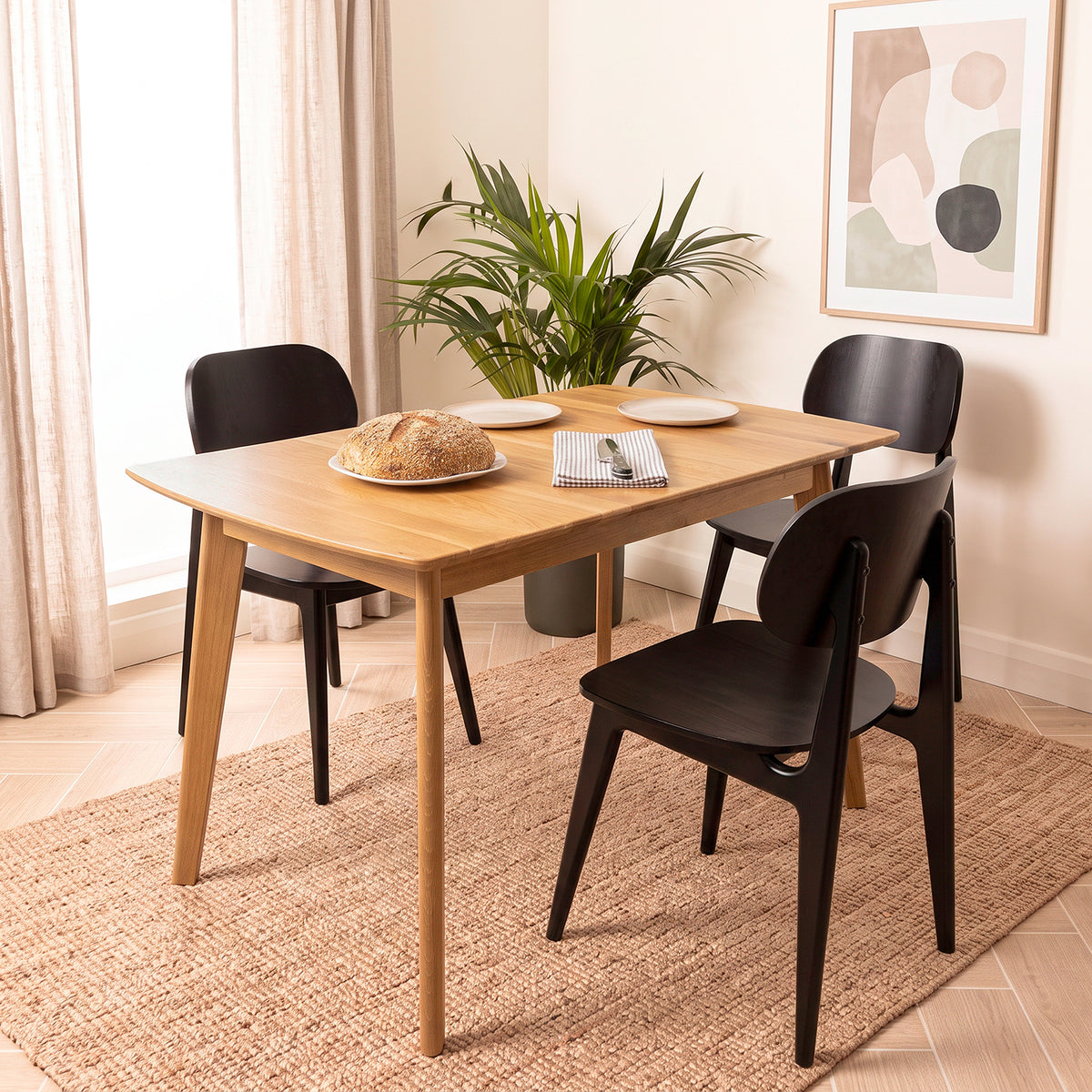 An oak wood rectangular dining table with tapered legs, surrounded by three black chairs, set on a woven jute rug in a bright, modern dining room.