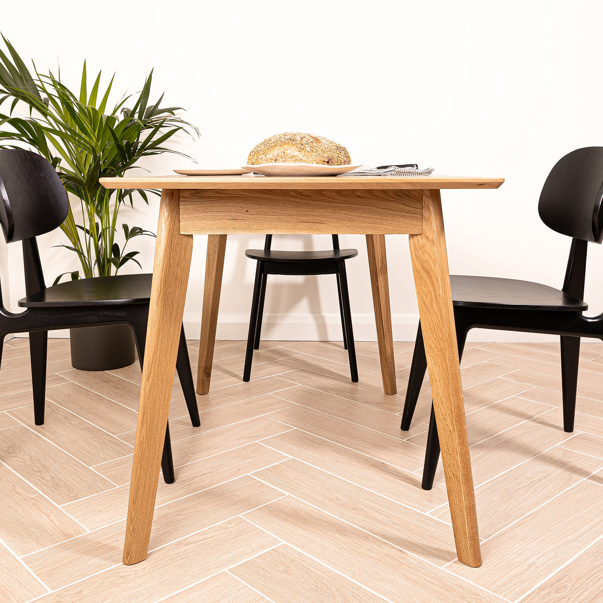 A light natural oak dining table with tapered legs, accompanied by three black chairs in a bright room with light wood herringbone flooring and a potted plant.