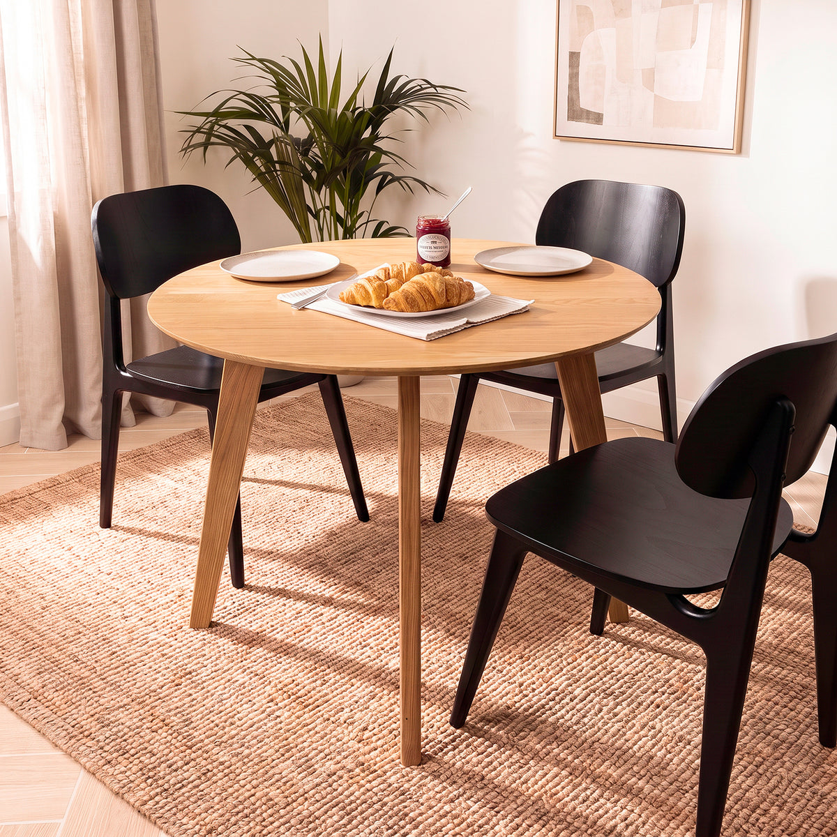 An Otti round oak wood dining table with three black chairs, styled with plates, croissants, a jar of jam, and a vase of flowers on a round woven jute rug in a bright room.