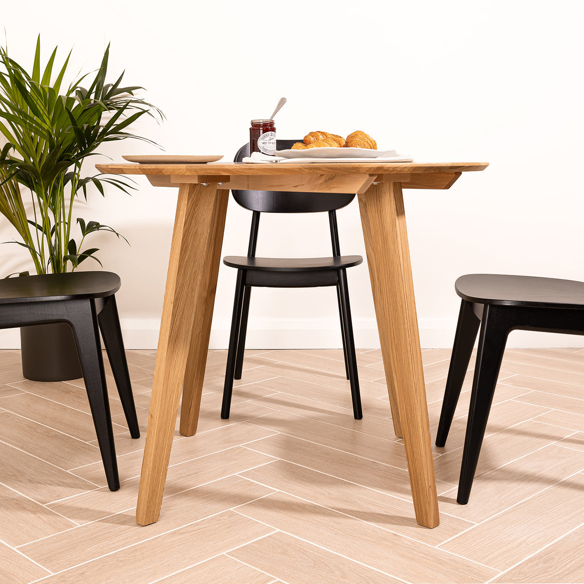 A light natural oak round dining table with three angled legs, shown in a bright room with black chairs, a potted plant, and a plate of pastries on top.