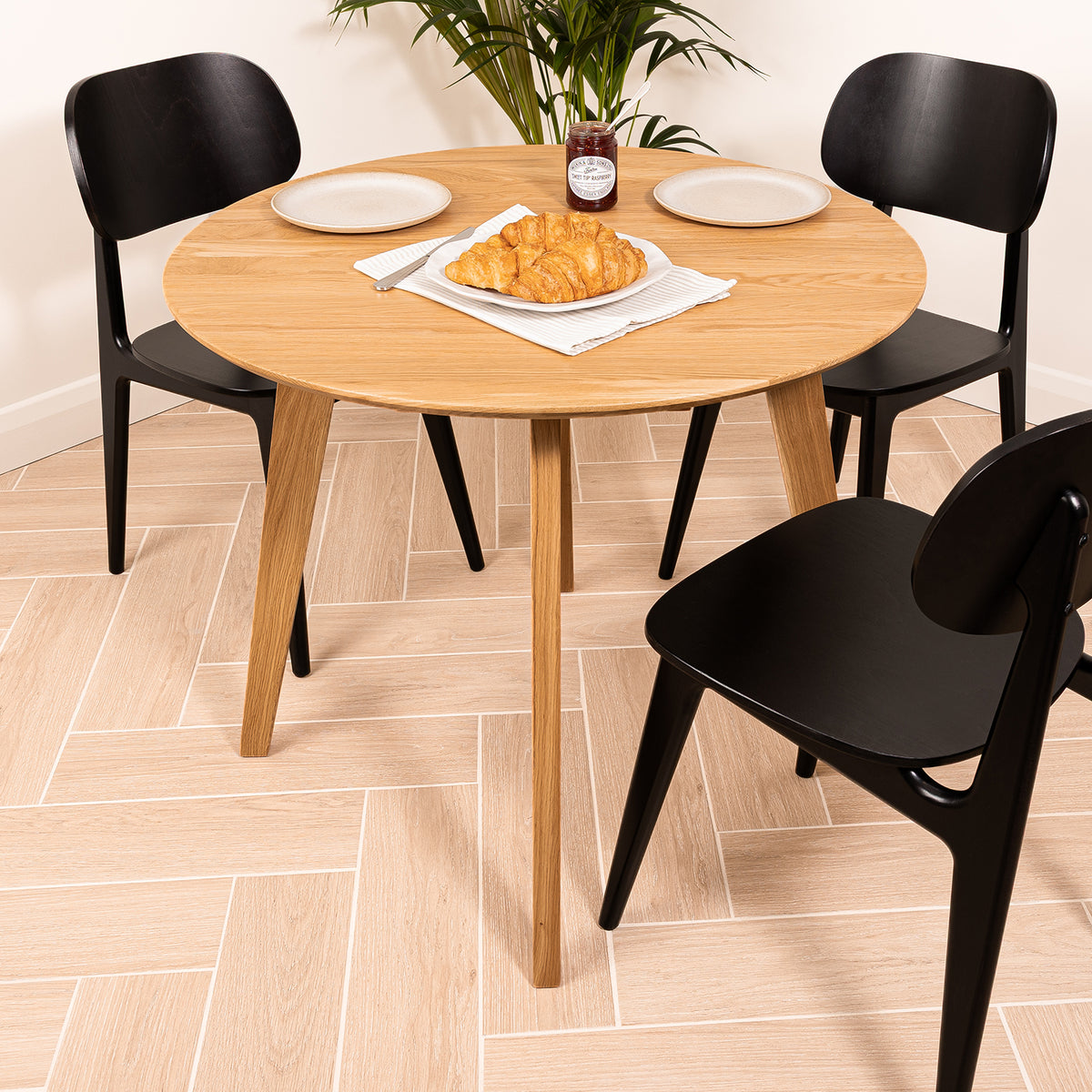 A Otti solid oak round dining table with three black chairs, styled with plates, a croissant, and a jar of jam on a light wood herringbone floor.