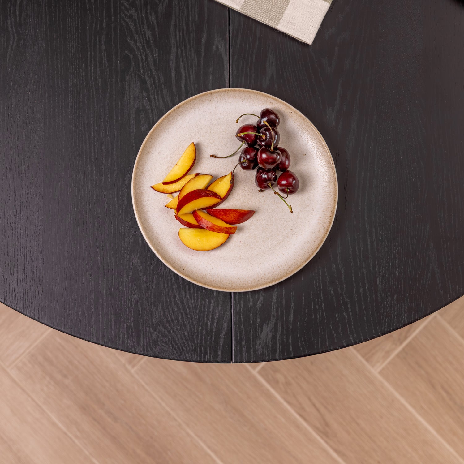 A round ceramic plate with a speckled beige glaze sits on a black oak wood extendable table, styled with sliced apples and a cluster of dark red cherries. The table is set against a light wood floor.