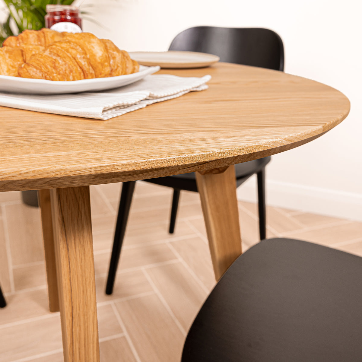 A solid oak round dining table with tapered legs, shown in a home dining setting with black chairs, a croissant on a plate, a jam jar blurred in the background, and a folded white napkin.
