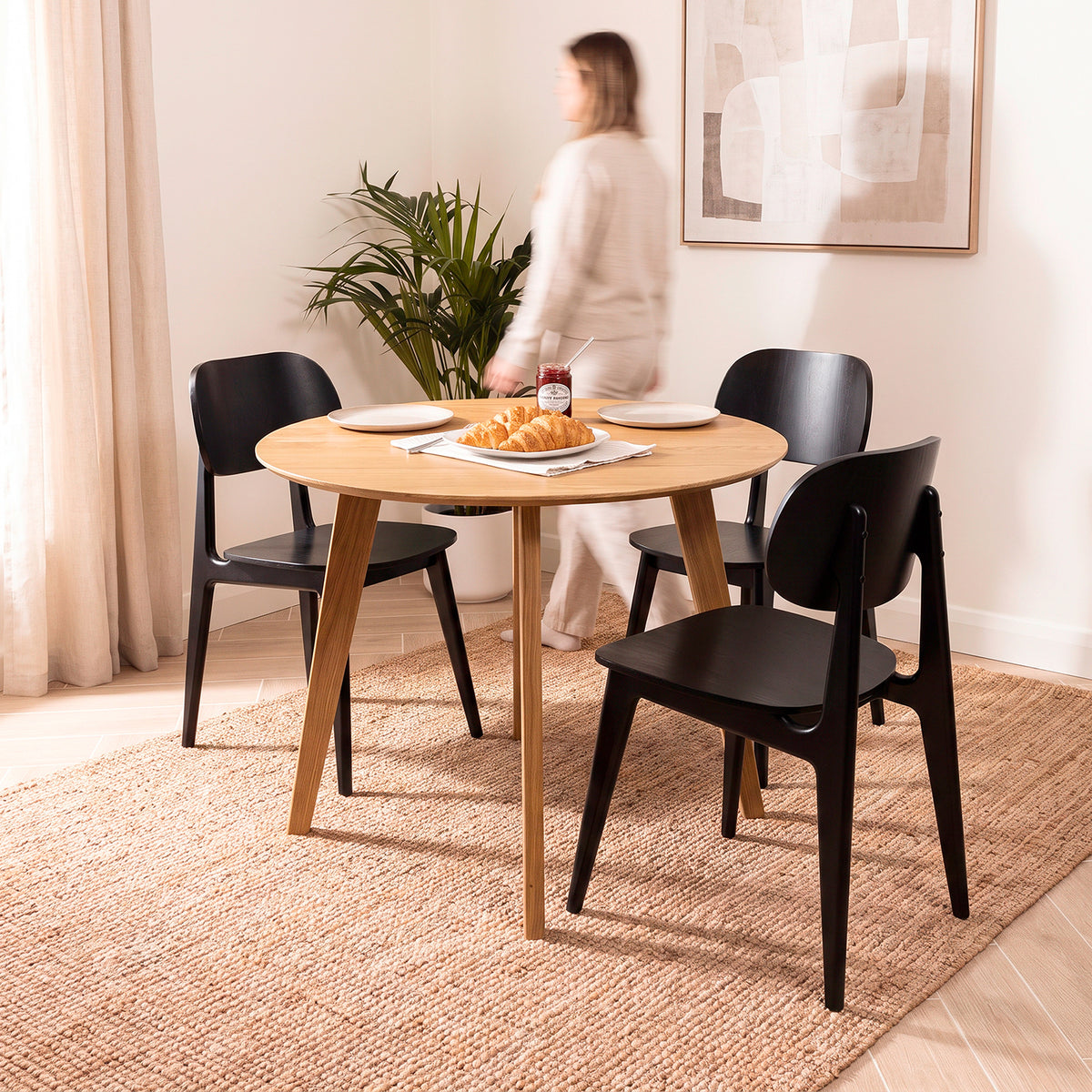 A round light oak wood dining table with two black chairs in a bright room, styled with plates, a croissants, and a potted plant. A woman is blurred in the background near a window.
