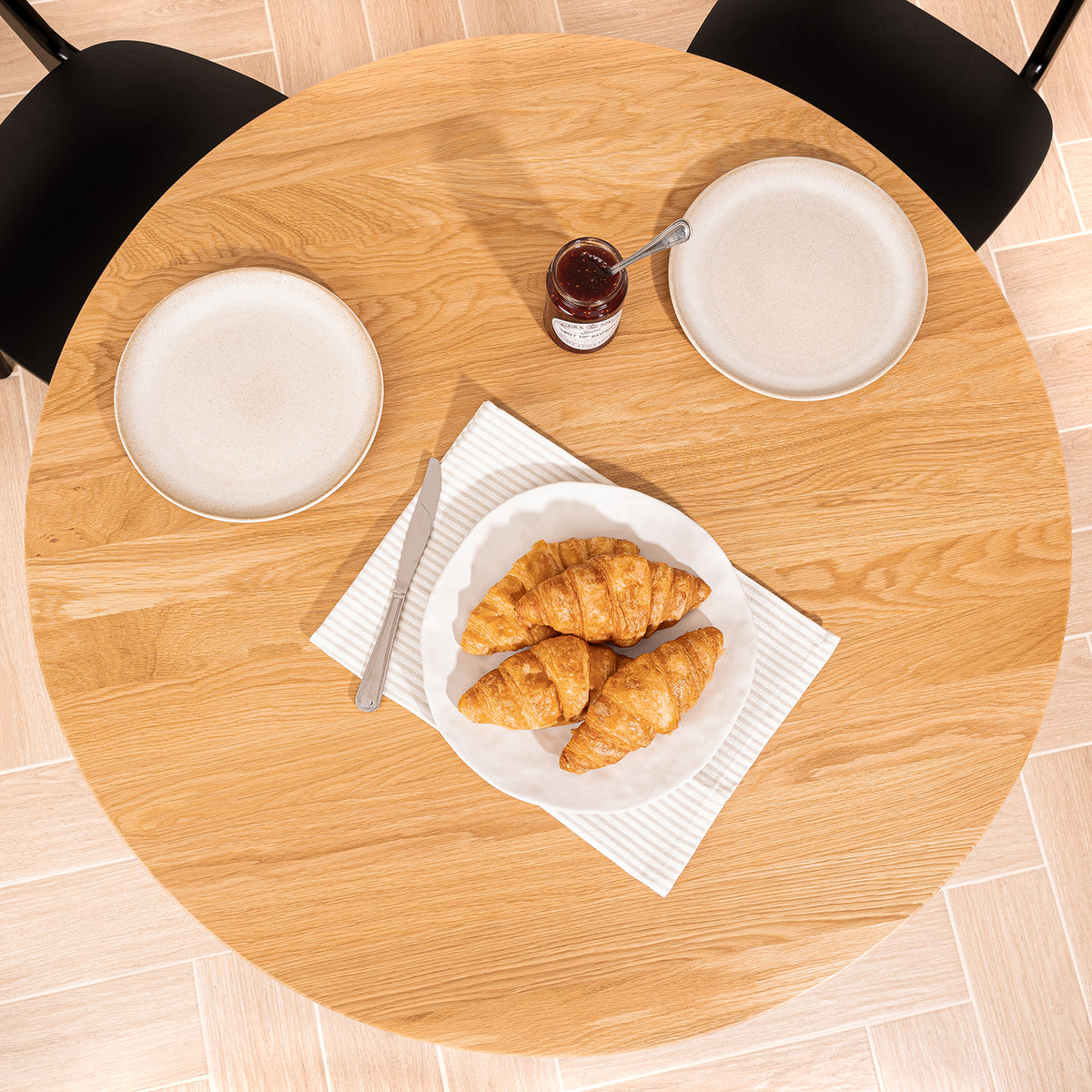 Top-down view of a round light oak dining table with black chairs, set with white plates, a small jar of jam, a knife, and a plate of four croissants on a white napkin.