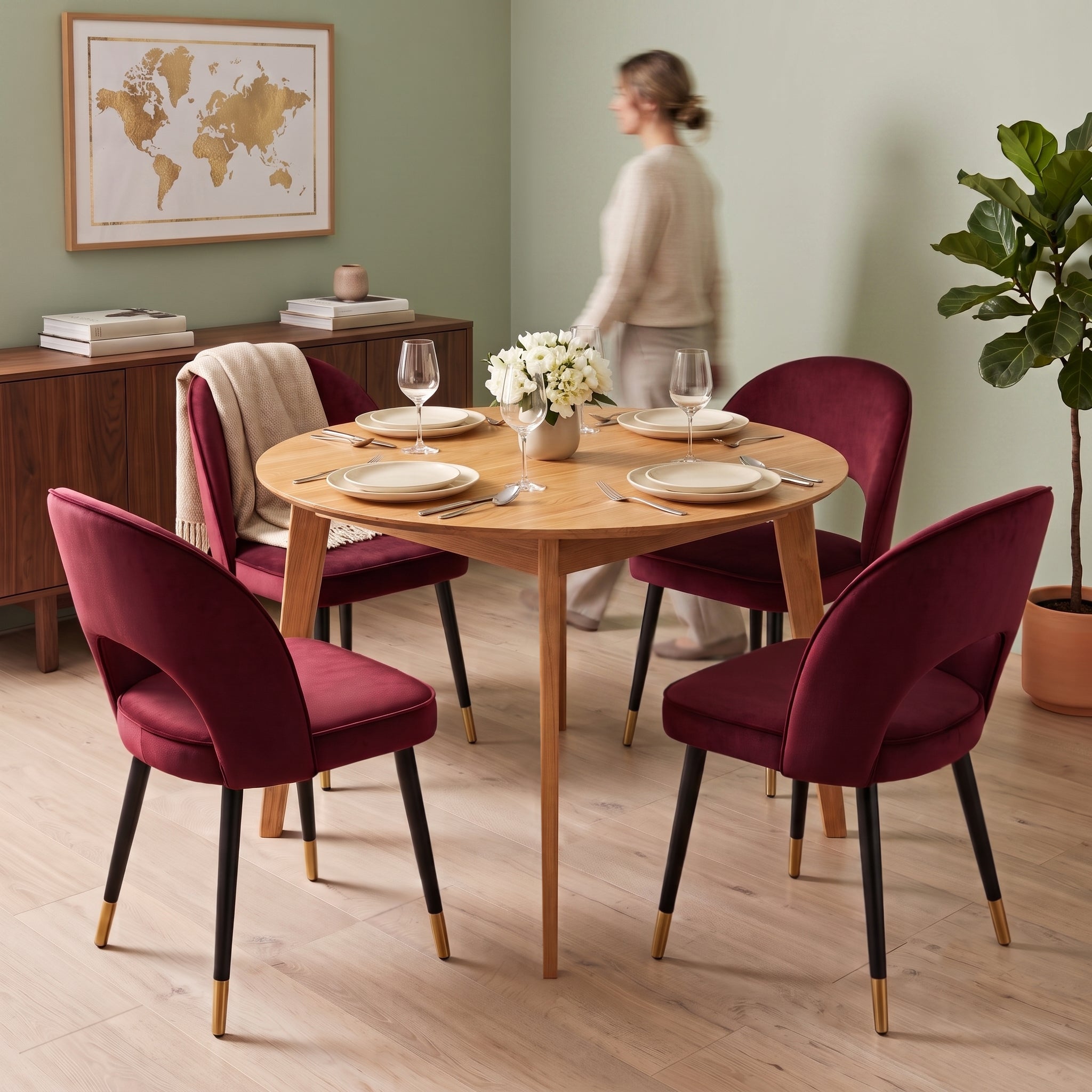 A round light wood dining table surrounded by four deep Oxblood velvet chairs with black legs and gold-tone tips, set in a styled dining room with a blurred woman walking in the background.