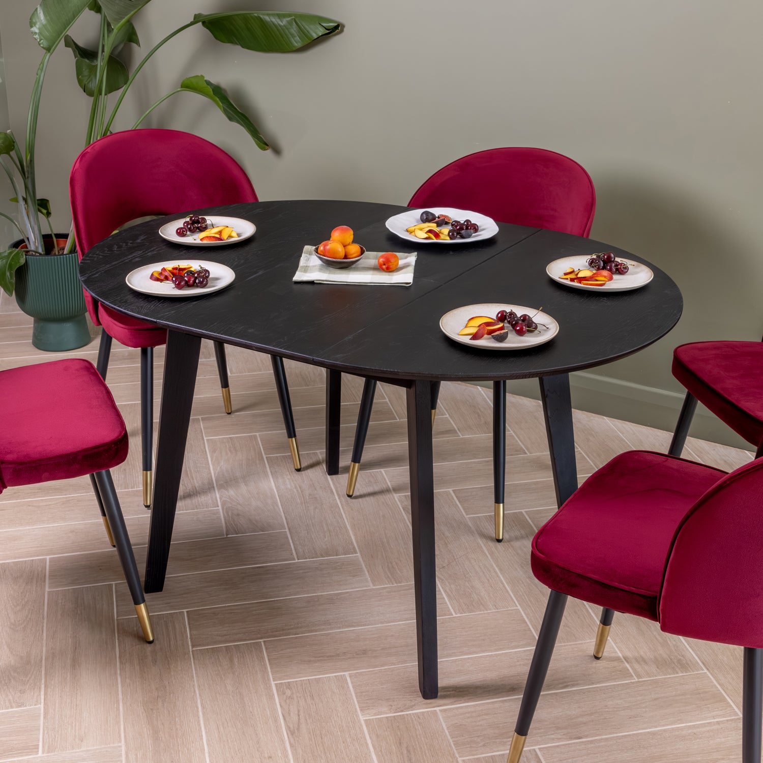 Round black dining table that has been extended with five oxblood velvet chairs, styled in a dining room setting with plates of fruit and a small centerpiece on a light wood floor. |  1375mm (extended)