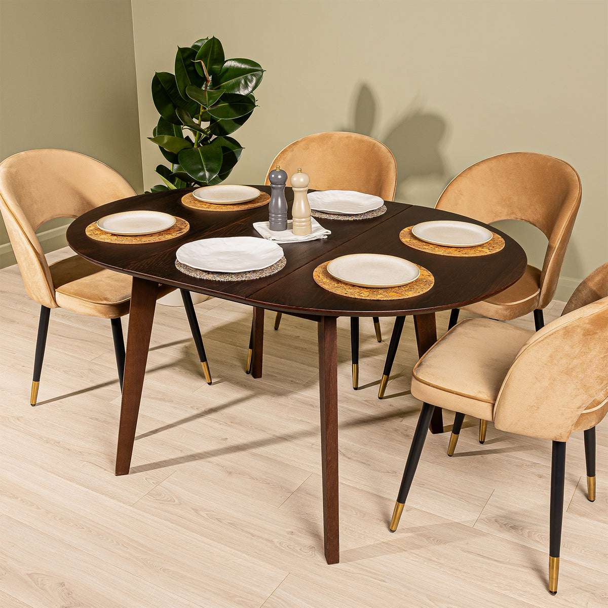 An extended round oak dining table in walnut finish with four Corin velvet dining chairs in a caramel finish, styled with plates and a plant in a modern dining room setting.