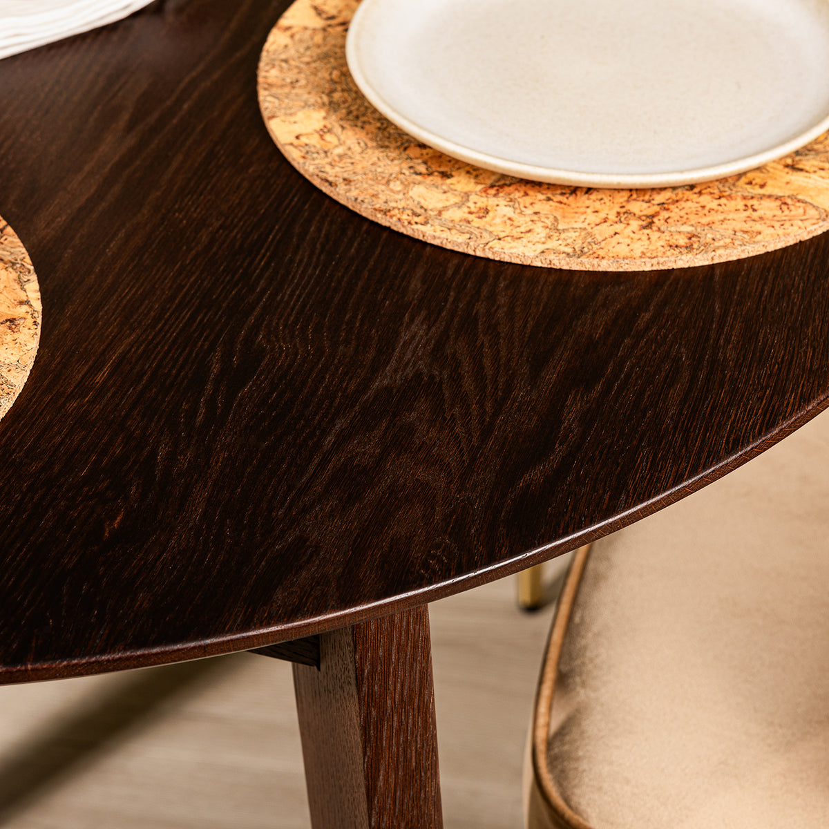 Close-up of a walnut finish oak wooden extendable dining table with a circular cork placemat and a white plate on top, with part of a caramel velvet chair visible.
