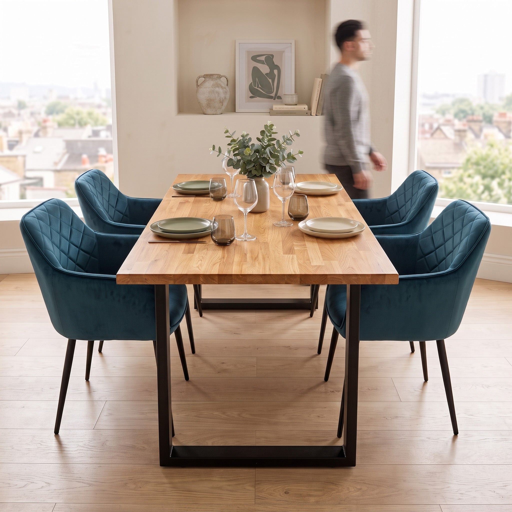 A modern dining room scene with a rectangular solid oak dining table and four bluevelvet chairs with black metal legs. A person is blurred in the background near large windows with an outdoor view.