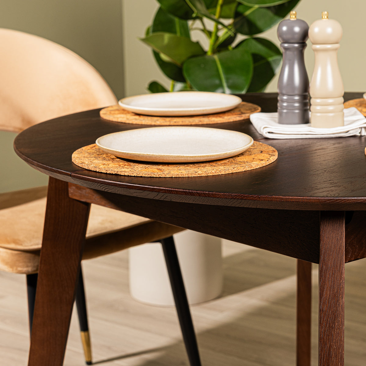 A walnut effect oak dining table set with round cork placemats, white ceramic plates, a folded white napkin, and two salt and pepper mills, with a Corin caramel velvet upholstered chair and a green plant in the background.